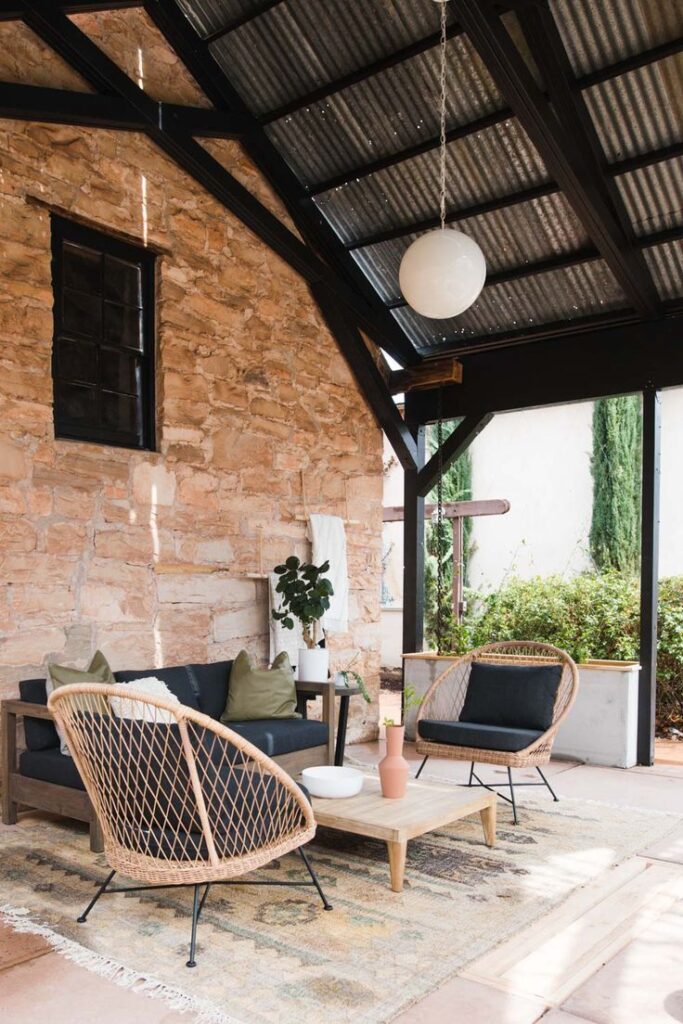 Cozy patio with wicker chairs, rustic stone wall, black sofa, and pendant light under slatted roof. Ideal for relaxation.