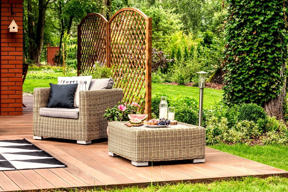 Outdoor patio with wicker chair, table, and snacks on a wooden deck, surrounded by lush greenery and lattice screen.