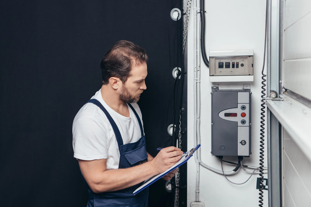 Electrician checking control panel with clipboard, wearing overalls and standing in a utility room.