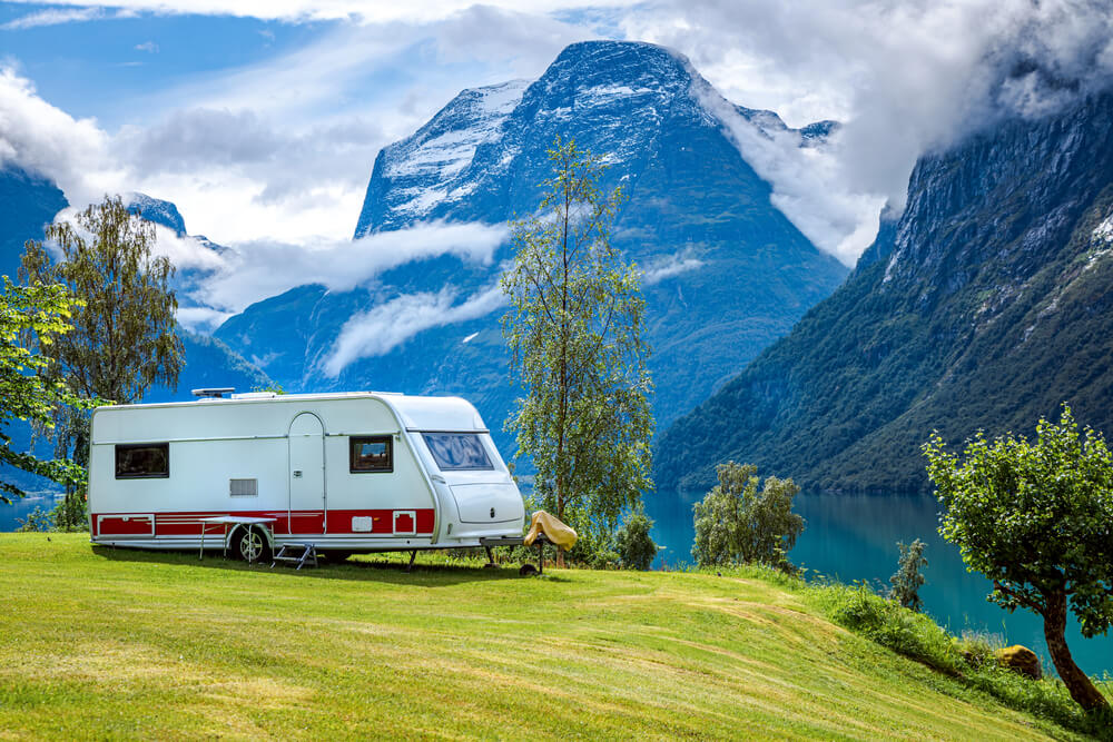 Caravan parked in a lush green landscape with scenic mountains and a lake in the background.