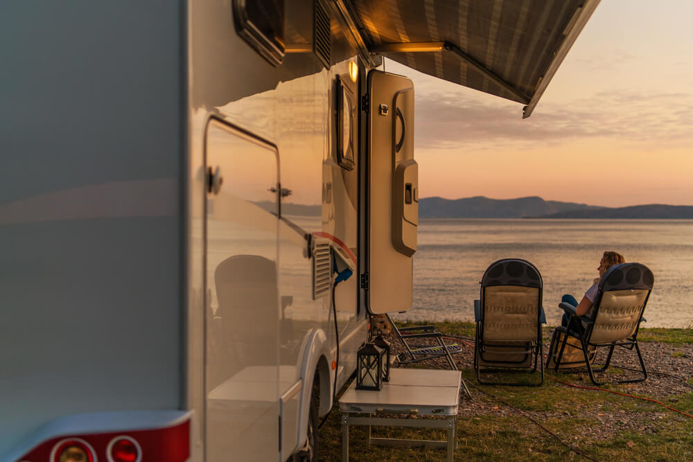 RV camper by a serene lake at sunset with person relaxing in a chair. Peaceful outdoor travel scene.