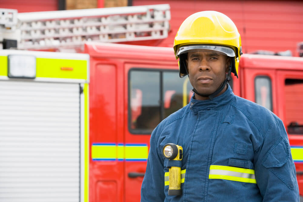 Firefighter in uniform standing in front of a red fire truck, wearing a helmet and safety gear.