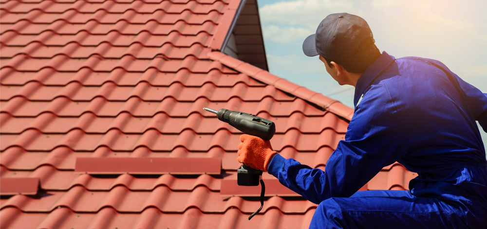 Roofer in blue uniform using a drill on a red tile roof under sunny skies.