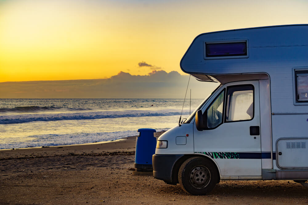 Camper van parked on a sandy beach at sunset, ocean waves and vibrant sky in the background.