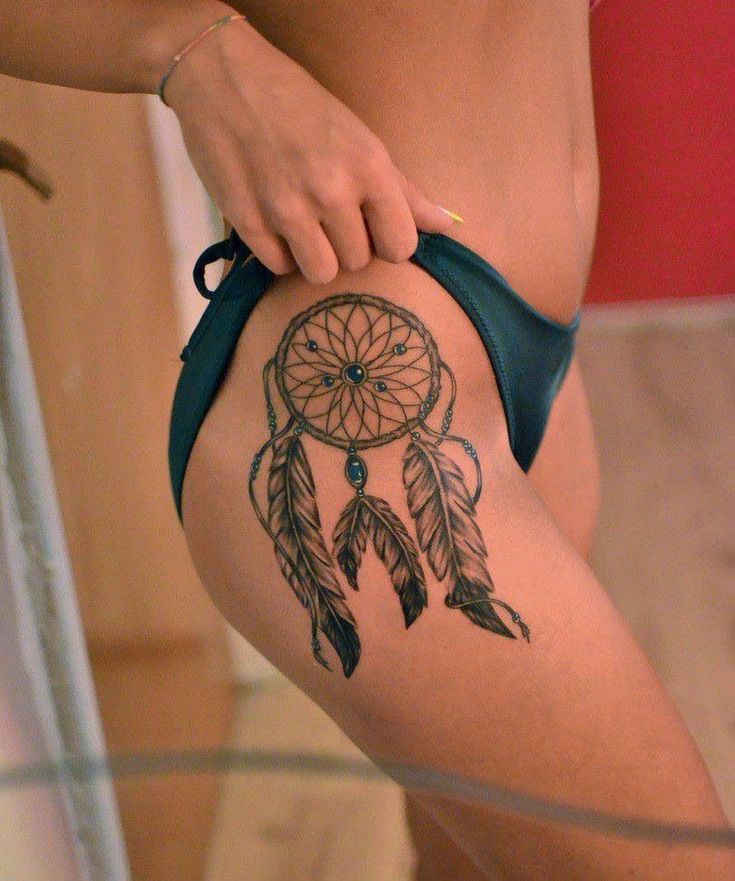 Dreamcatcher tattoo on woman's thigh in blue bikini, showcasing intricate feather design.