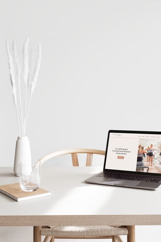 Minimalist workspace with laptop displaying website, white vase, glass, and chair on light-colored desk.