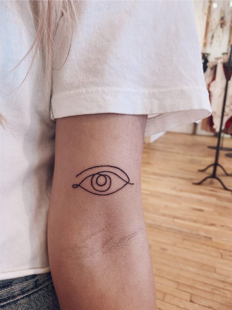 Minimalist eye tattoo on arm, visible from the side, with wooden floor background and a white shirt sleeve.