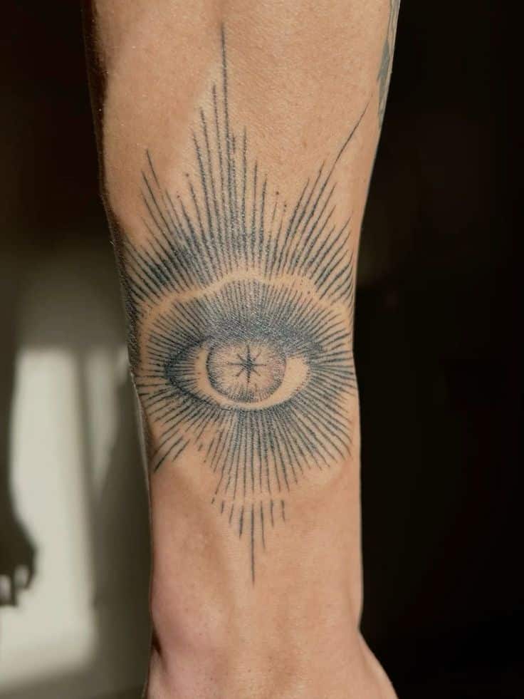 Intricate eye tattoo with sunburst rays on inner arm. Black ink design with symmetrical lines.