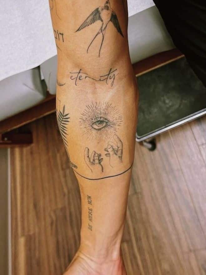 Arm with various tattoos including a bird, eye, hands, and the word eternity. Tattoos are on a wooden floor background.