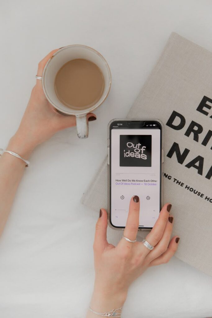 Person holding a coffee cup and smartphone with podcast playing, laid on a book. Cozy morning routine.