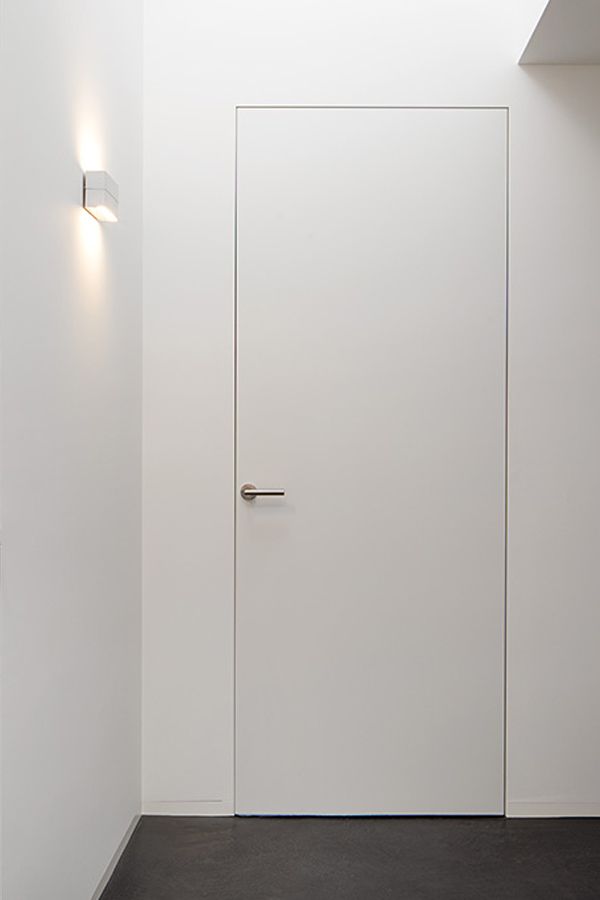 Minimalist white wall with flush door and modern handle, illuminated by a wall sconce, creating a sleek design.