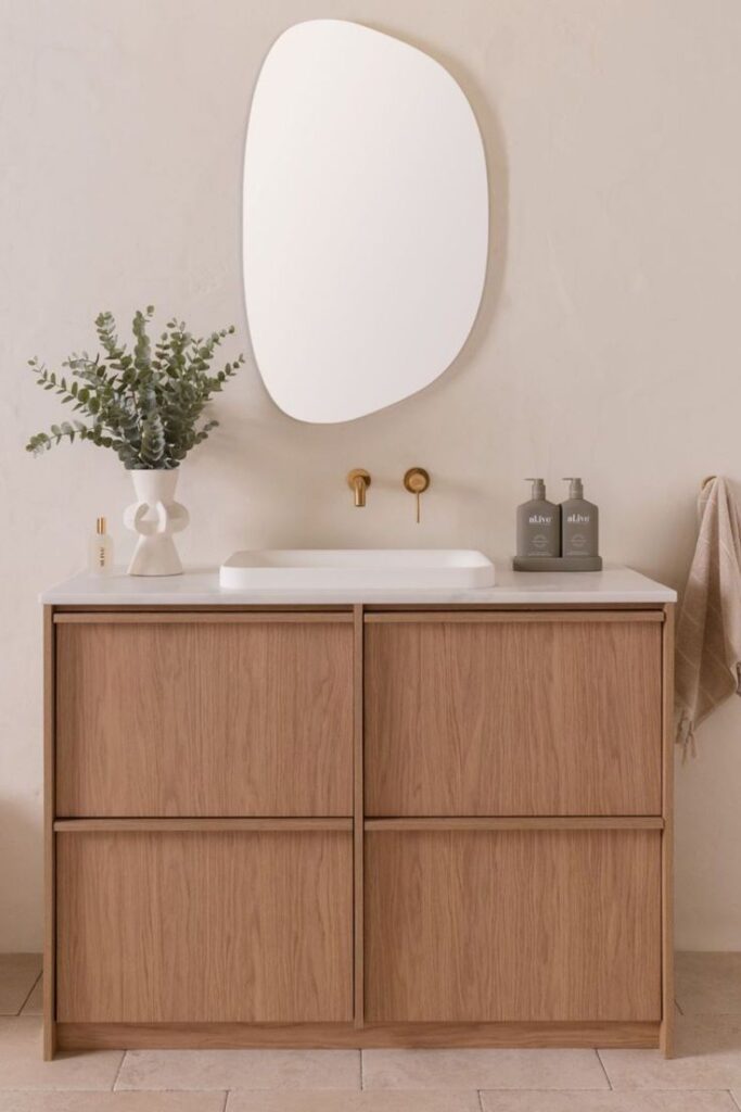 Modern bathroom with wooden vanity, abstract mirror, and stylish decor, featuring eucalyptus in a vase.