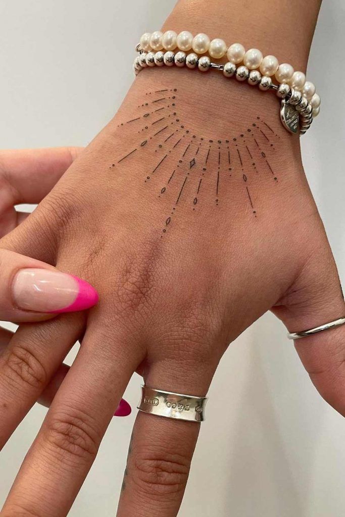 Hand with minimal sunburst tattoo, wearing pearl bracelets and silver rings, showcasing a pink-tipped manicure.