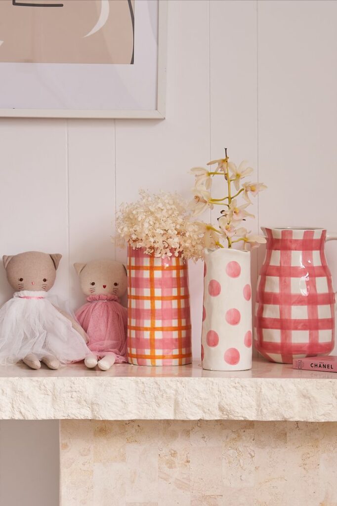 Chic shelf decor with plush cats, checkered vases, flowers, and Chanel book on a white background. Cozy home style.