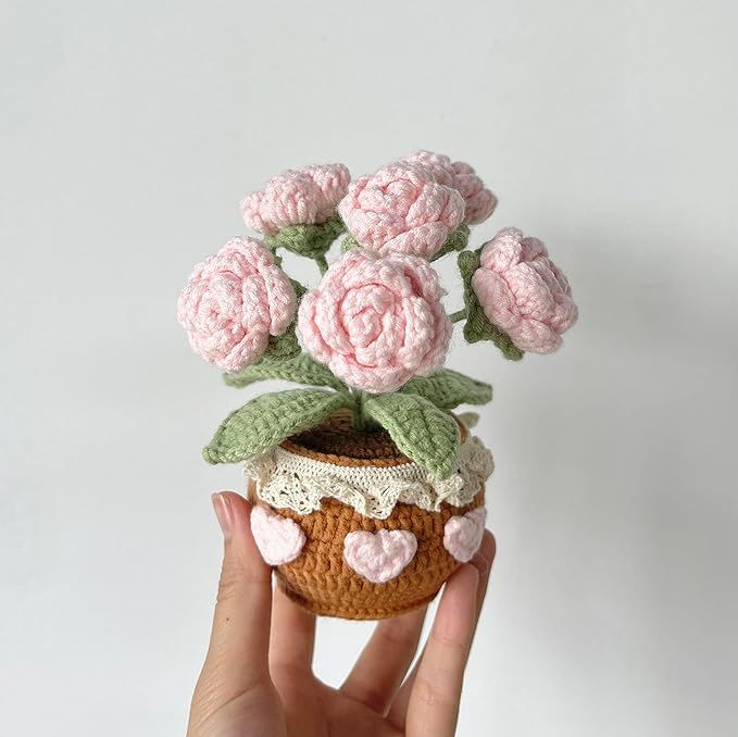 Hand holding a crocheted pink flower bouquet in a decorated pot with lace and heart accents.