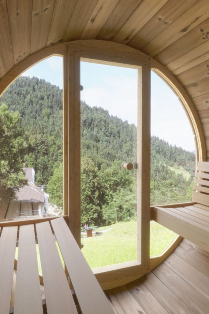 Cozy wooden sauna with large round window overlooking lush green forest and hills.