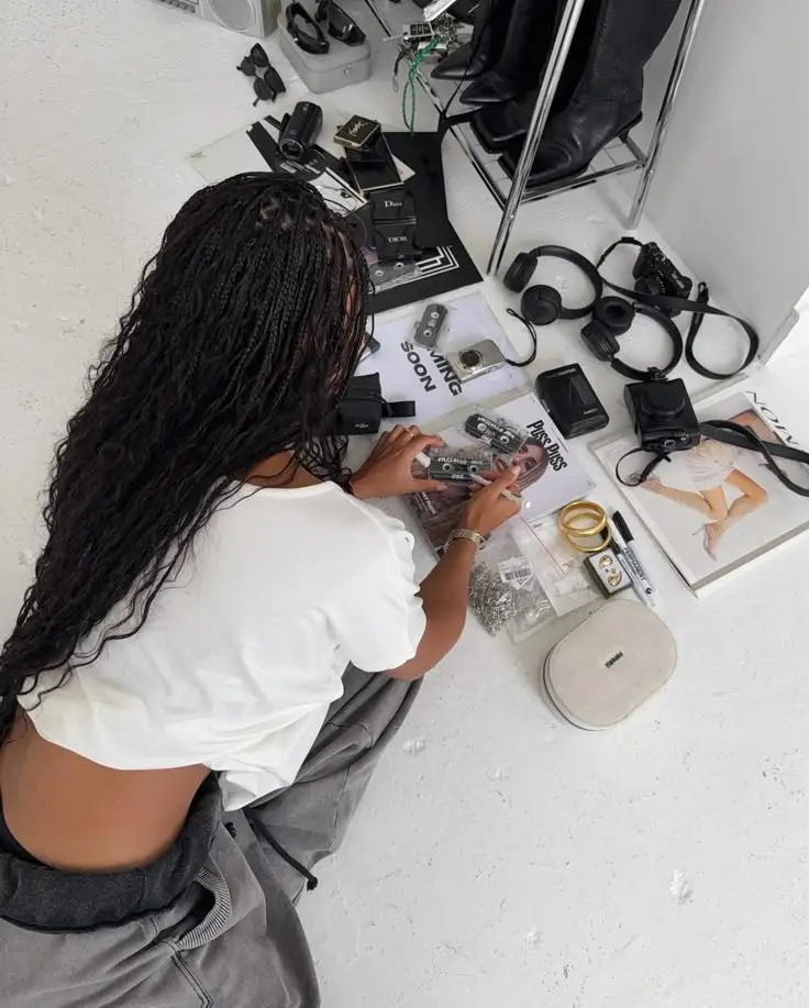 Person organizing gadgets, cameras, and magazines on a white floor, surrounded by tech accessories and fashion items.