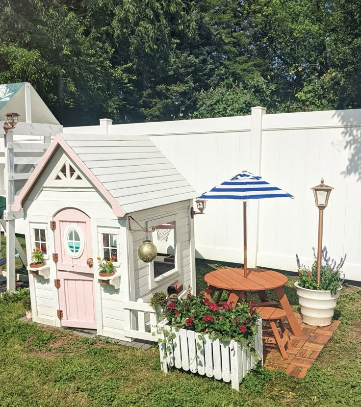 Charming playhouse with a picnic table, umbrella, and lush garden in a sunny backyard setting.