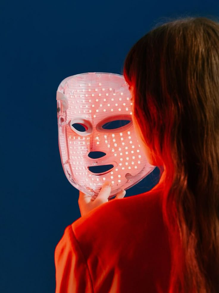 Person holding an LED face mask, glowing red light therapy for skincare, against a deep blue background.