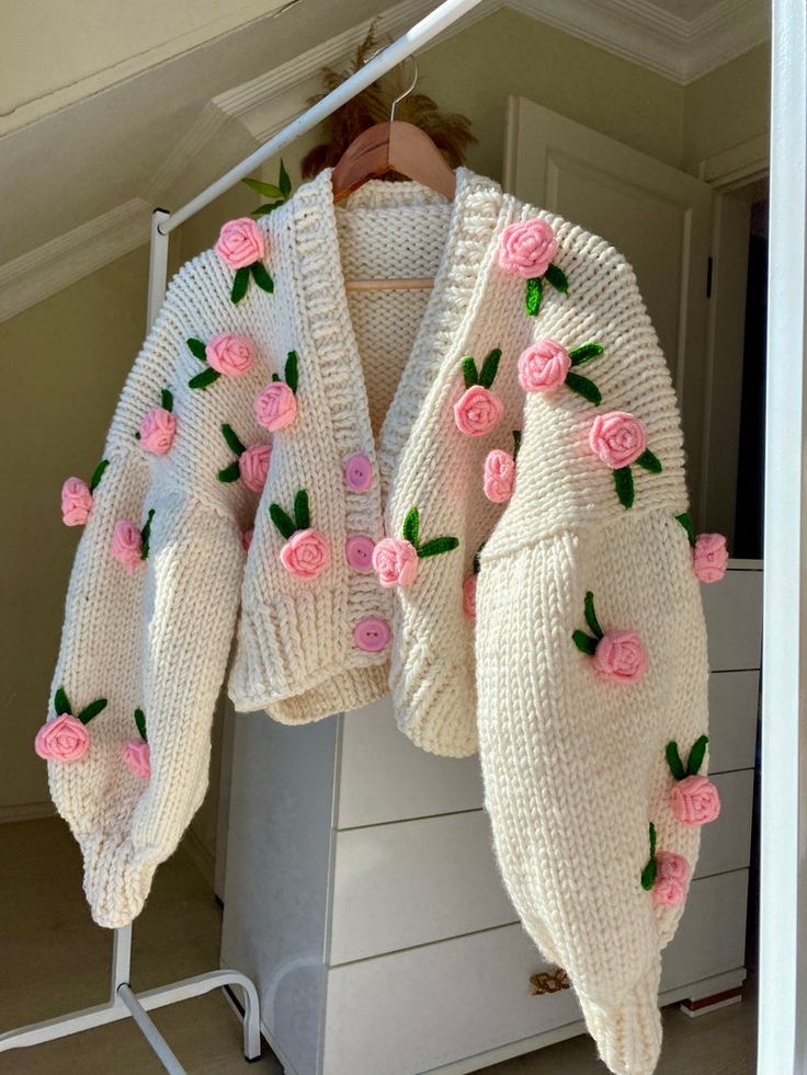 Cozy hand-knitted cream cardigan with pink rose details, hanging on a wooden hanger in a bright room.