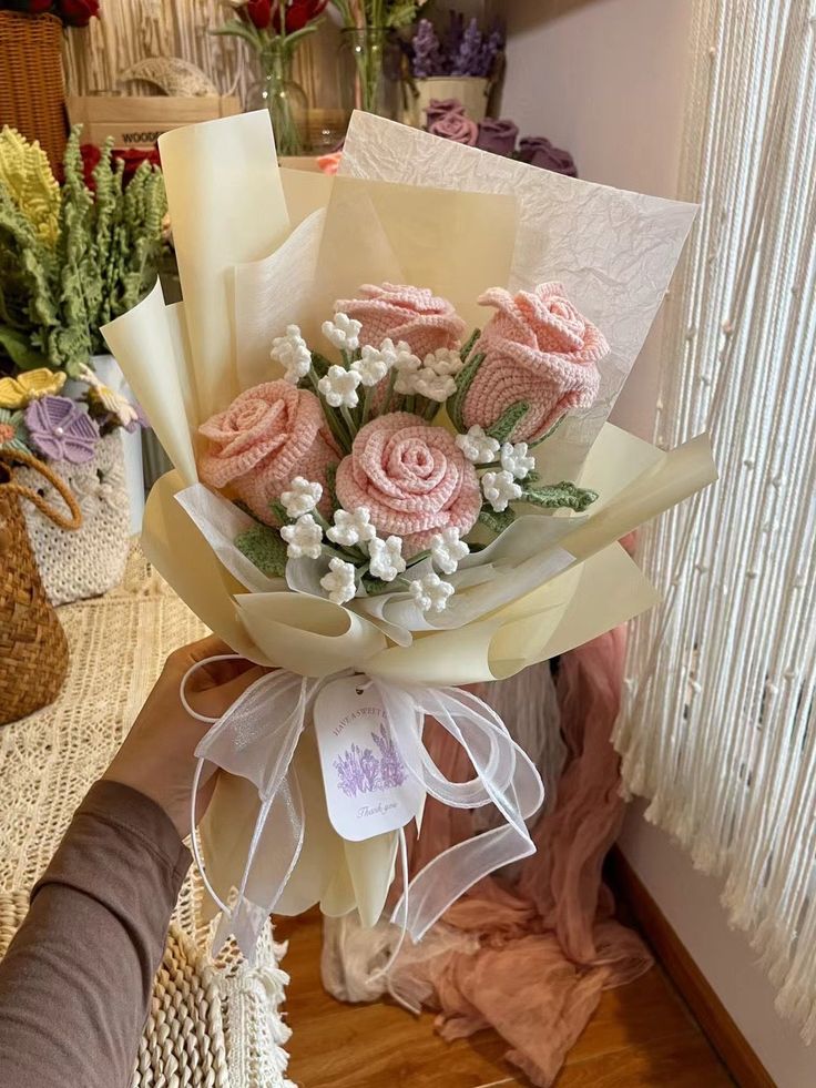 Crochet pink rose bouquet with white accents wrapped in paper, held in a cozy room setting.