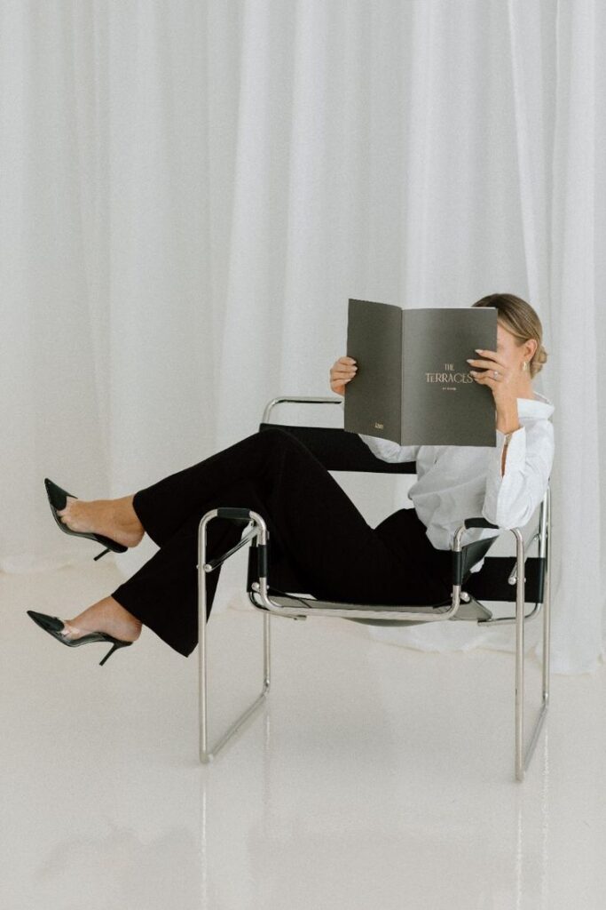 Person in black and white attire reading a book on a modern chair against a white background. Classy and minimalist.