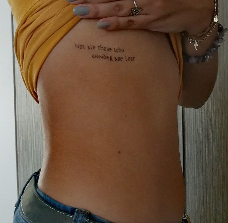 Side tattoo with phrase Not all those who wander are lost on a person's rib under a yellow top.