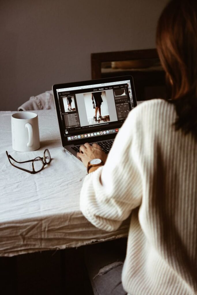 Person editing photos on a laptop at a table with coffee mug and glasses nearby.