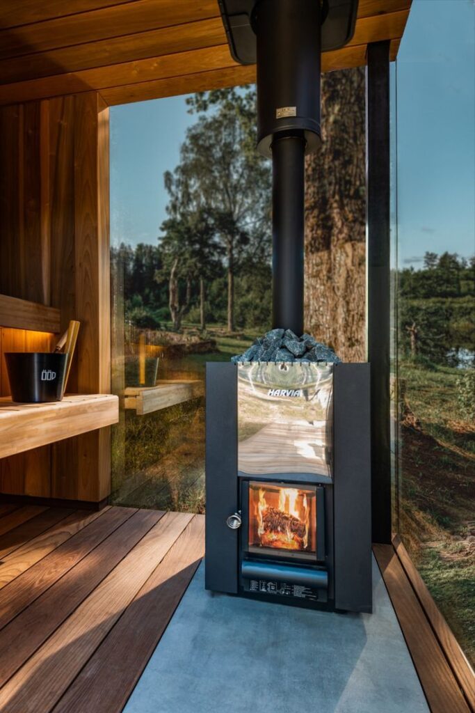 Modern sauna with a lit wood-burning stove, surrounded by glass walls showcasing a scenic outdoor view.