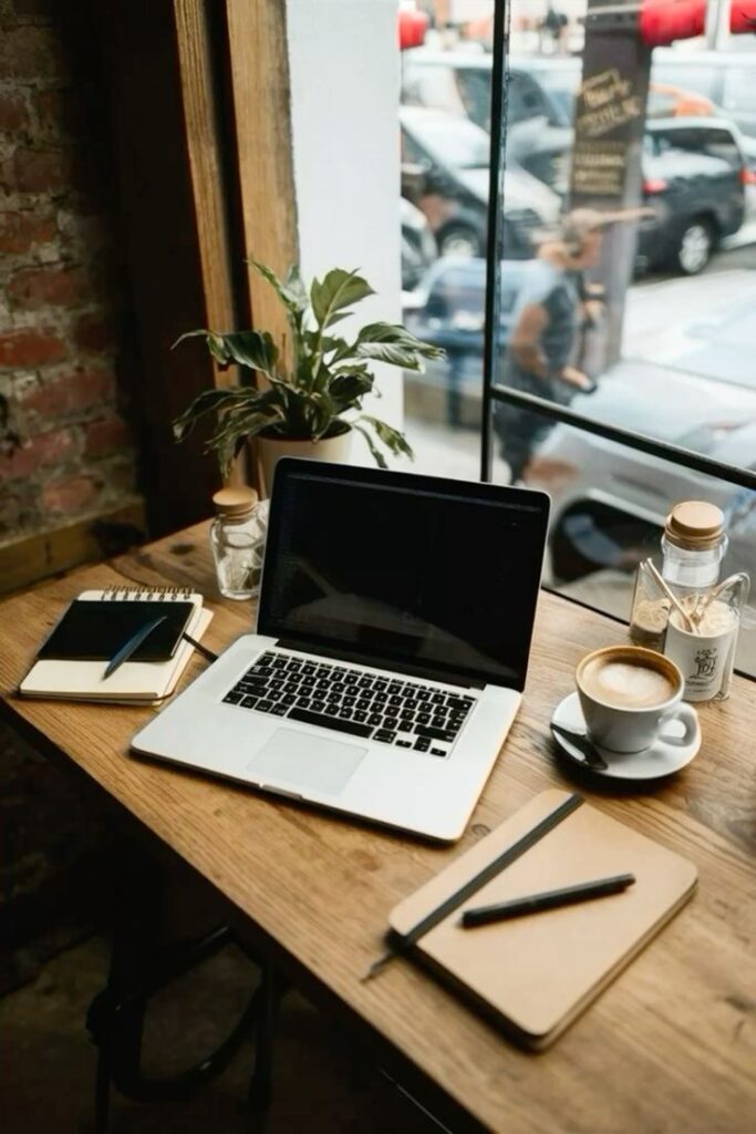 Cozy cafe workspace with laptop, notebook, coffee, and potted plant by a sunny window. Perfect for productivity.