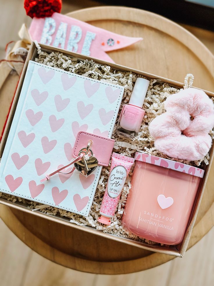 Gift box with pink heart-themed items, including a journal, candle, lip balm, nail polish, and scrunchie.