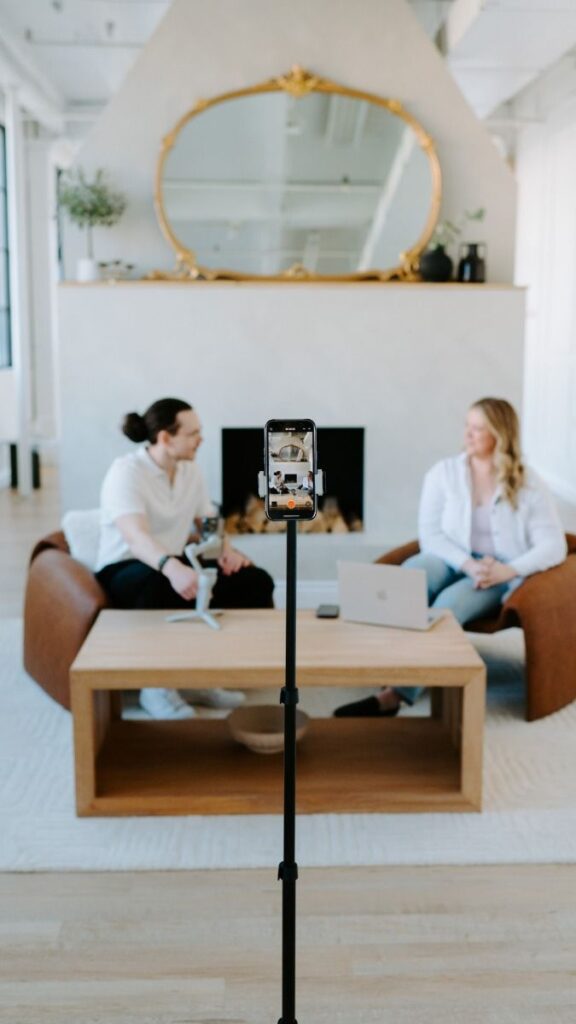 Recording a podcast: two people talk on a sofa, phone on tripod capturing the session, cozy modern setting.