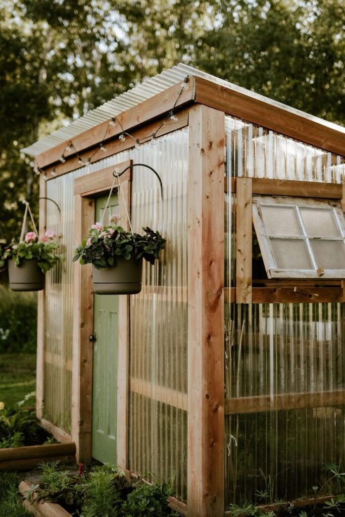 Rustic wooden greenhouse with hanging flower baskets and open window, surrounded by greenery.