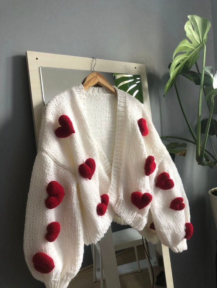 Cozy white knit cardigan with red heart accents hung on a mirror in a stylish room.