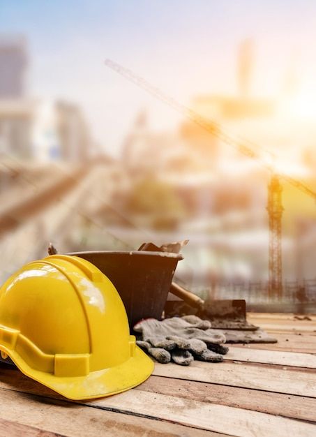 Yellow safety helmet and gloves on construction site with blurred cranes and sunset in background.