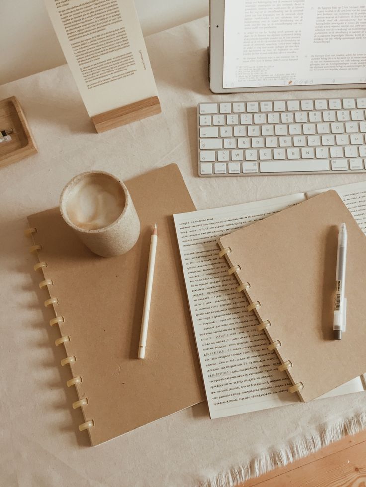 Minimalist workspace with a tablet, keyboard, spiral notebooks, a coffee cup, and a pencil on a light tablecloth.