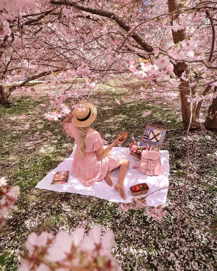 Woman enjoying a picnic under cherry blossoms, wearing a pink dress and hat, surrounded by spring blossoms and snacks.