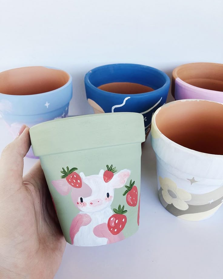 Hand-painted flower pots with cute designs, including a strawberry-themed animal, displayed creatively.