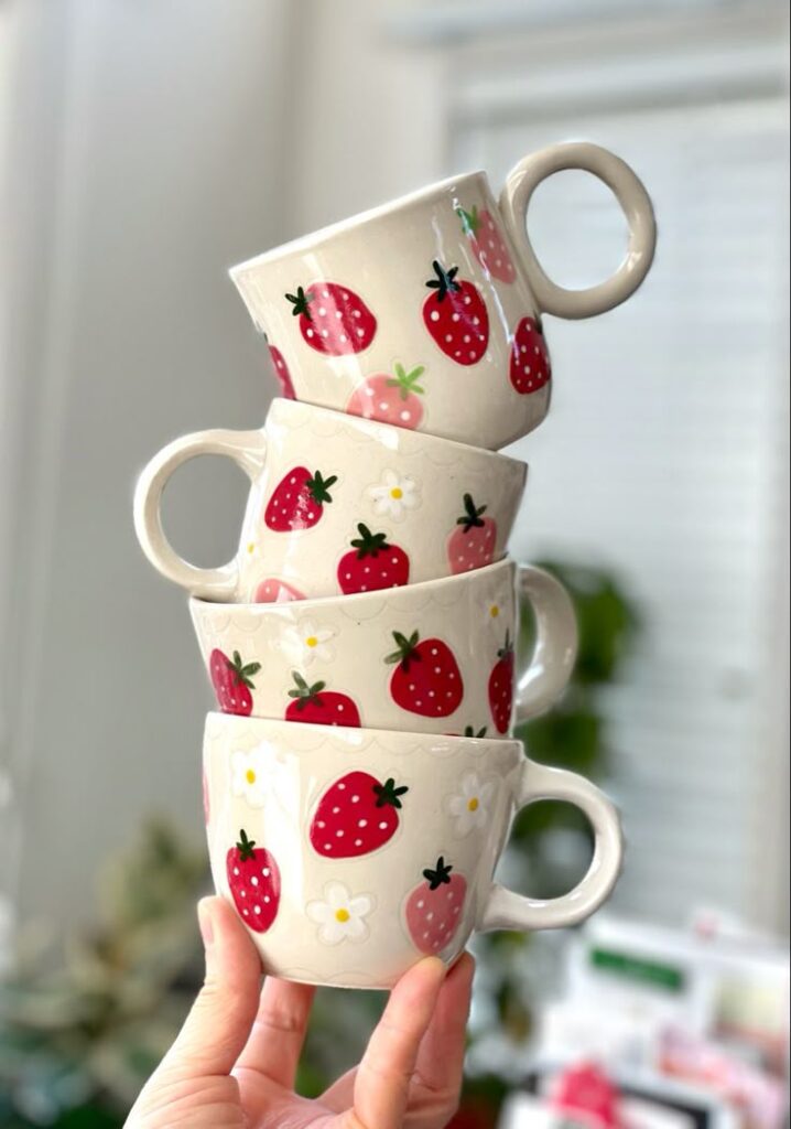 Stacked ceramic mugs with strawberry designs held in hand against blurred background.