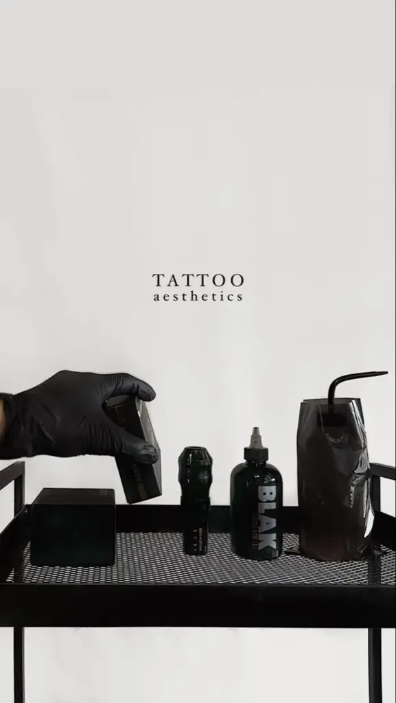 Tattoo supplies on a black tray including ink bottle and glove-hand holding a device, with Tattoo Aesthetics text.