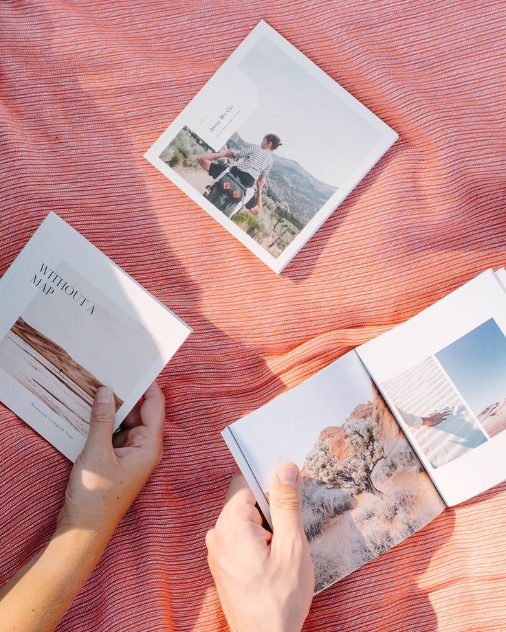 Hands holding travel photo books on an orange blanket, featuring landscapes and adventure themes.