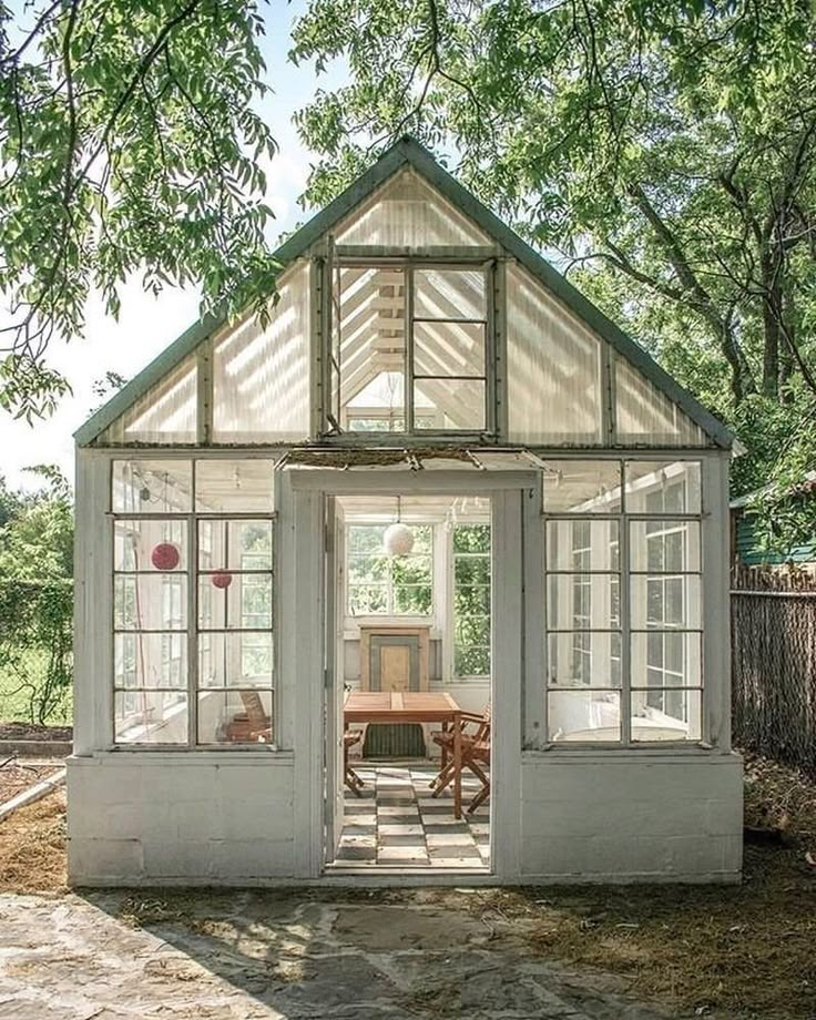 Charming rustic greenhouse with large windows surrounded by lush greenery, featuring a cozy interior and wooden chairs.