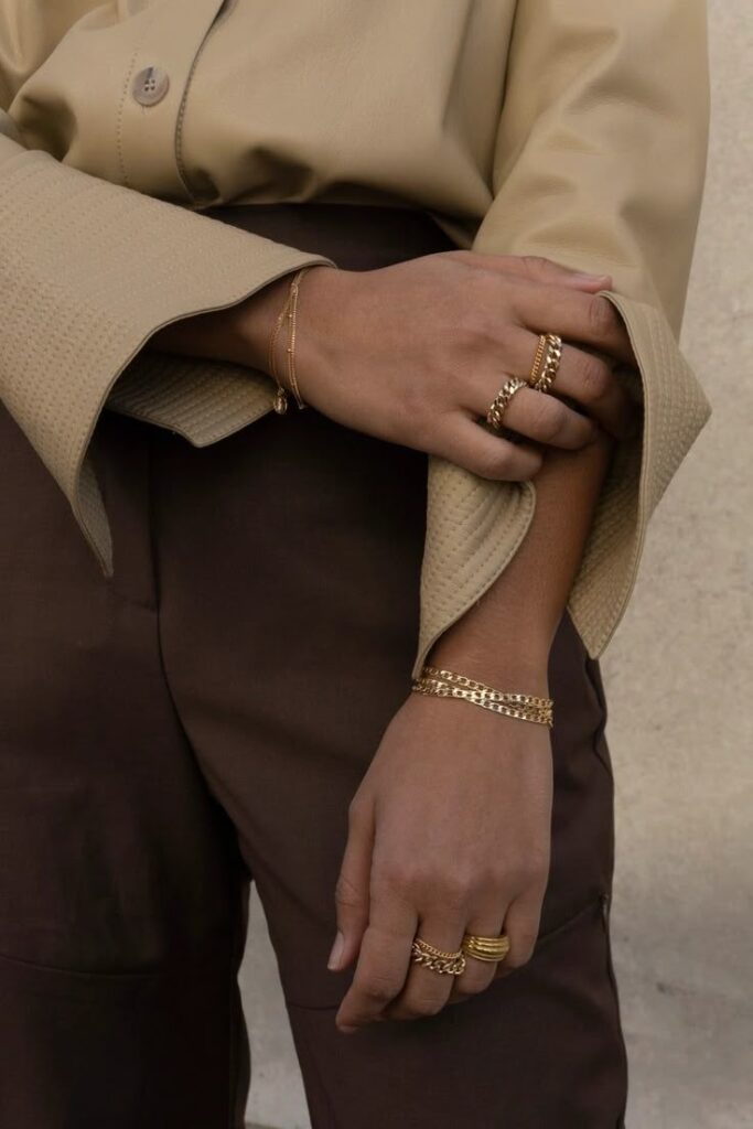 Elegant gold jewelry on person wearing beige top and brown pants, featuring rings and bracelets.