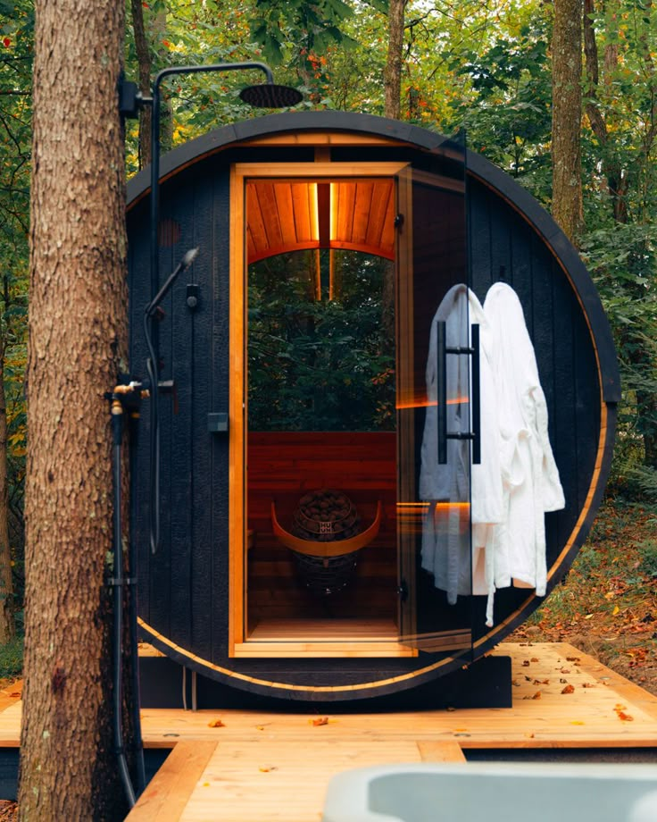 Round outdoor sauna in a forest setting with open door and robes hanging on the side.