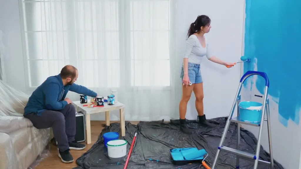 Couple painting a room with blue paint, creating a fresh look with rollers and tools on a plastic-covered floor.
