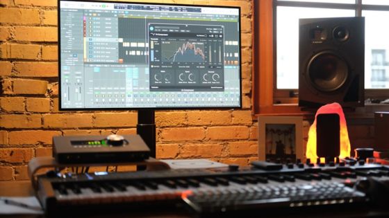 Music production studio with keyboard, monitor displaying audio software, speakers, and brick wall background.