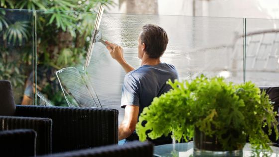 Man cleaning glass window on a sunny patio surrounded by greenery and outdoor furniture.