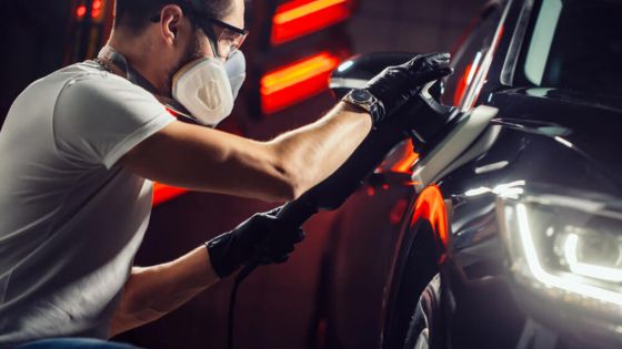 Person polishing a car with a buffer tool, wearing protective gear in a garage setting.