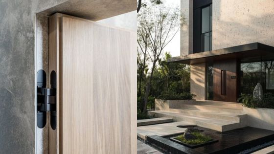 Modern house entrance with sleek wooden door and concealed hinges, surrounded by lush greenery and minimalist architecture.