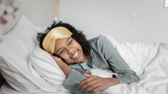 Smiling person relaxing in bed with a comfy pillow and eye mask, promoting restful sleep and relaxation.